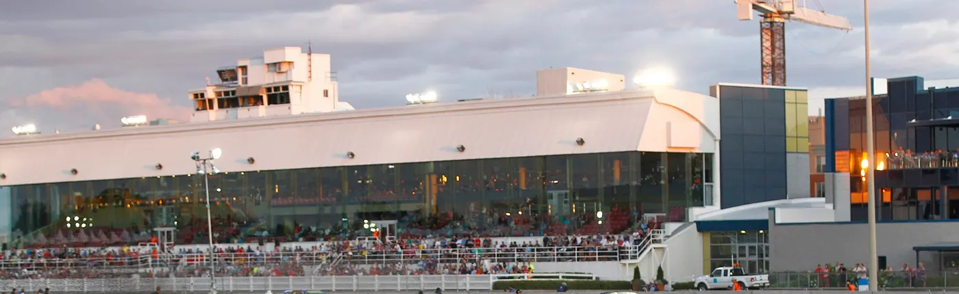 Photo : la tribune vitrée de l'Hippodrome de Trois-Rivières au crépuscule, des spectateurs le long de la clôture
